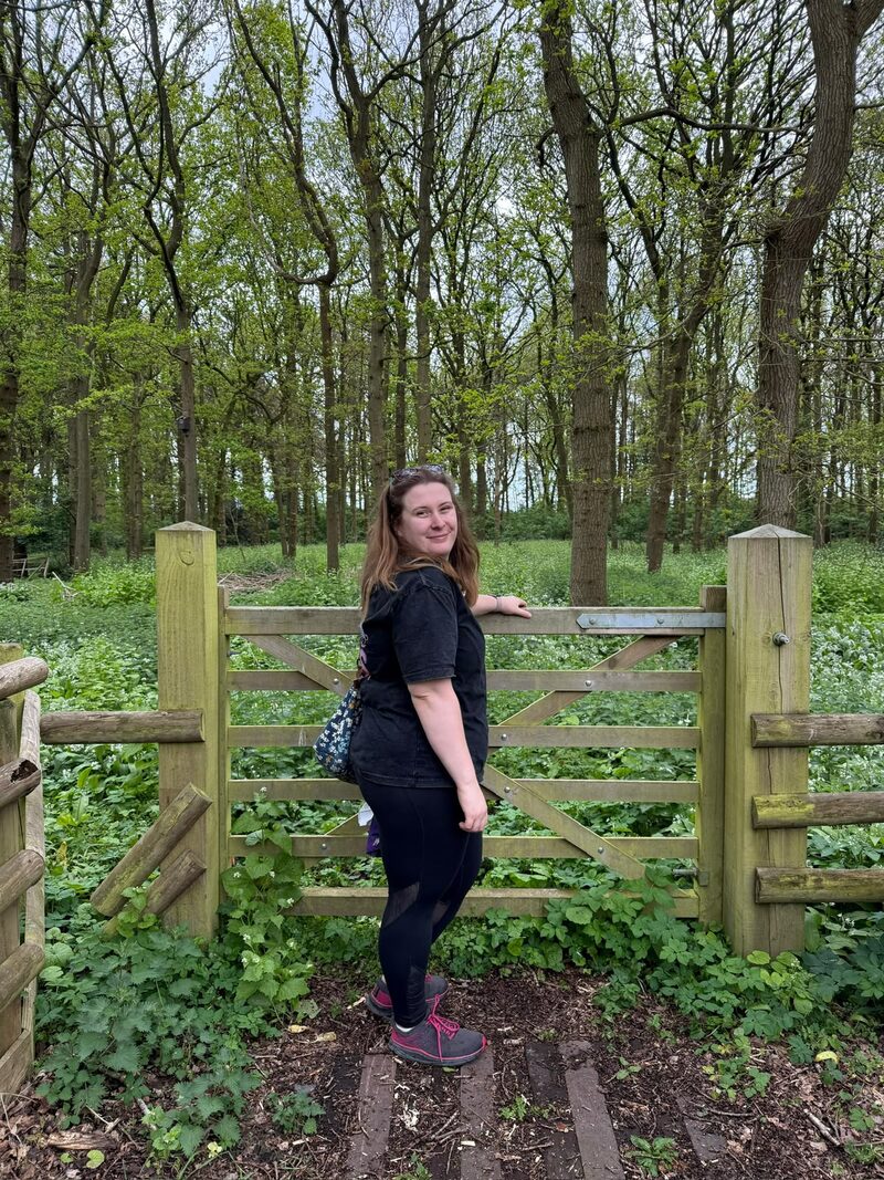 Emily standing at a wooden gate in a woodland setting, smiling and looking towards the camera