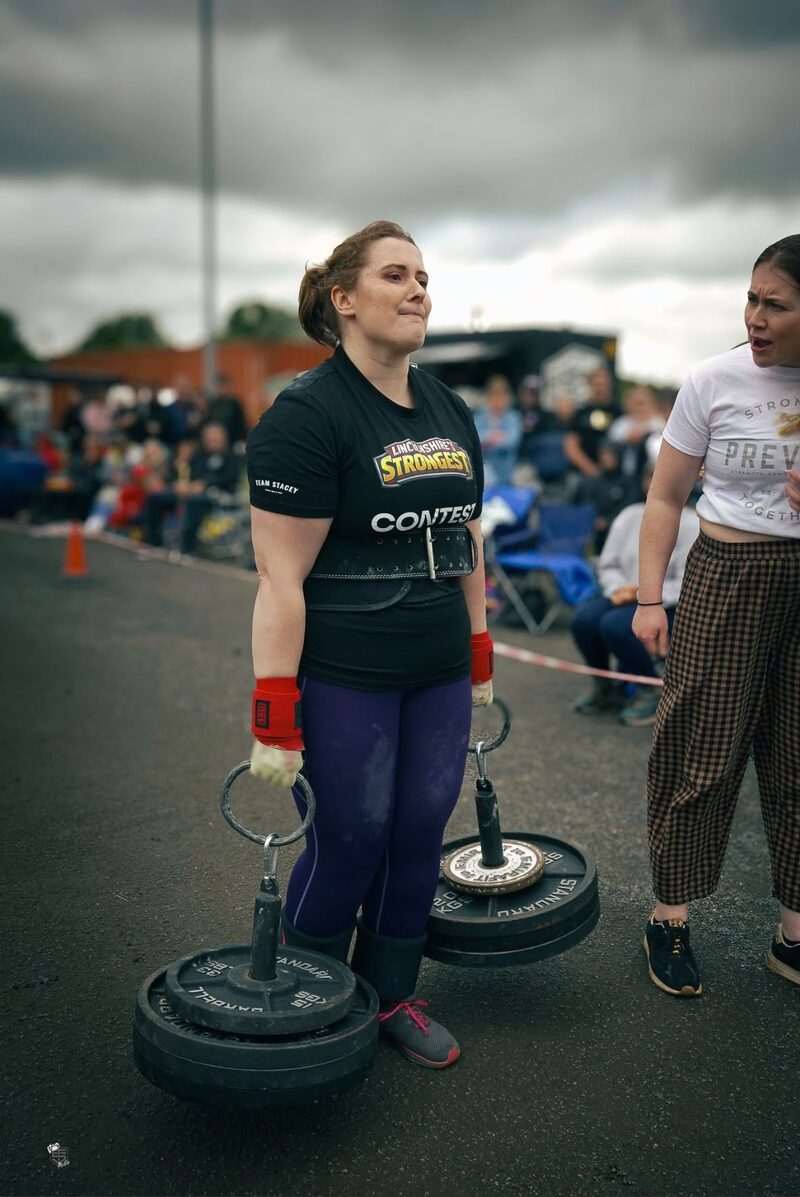 Emily competing at a strongwoman event, wearing a Lincolnshire's Strongest Contest shirt and lifting belt, mid-carry with heavy weight plates on metal handles